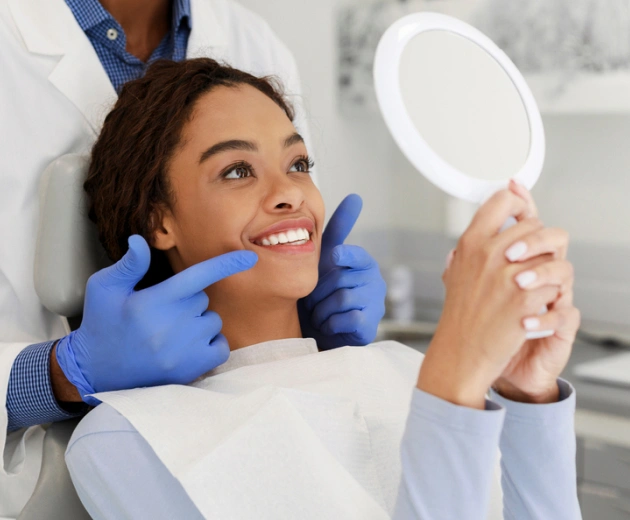 Patient examining teeth in mirror at dental office with dentist in blue gloves.