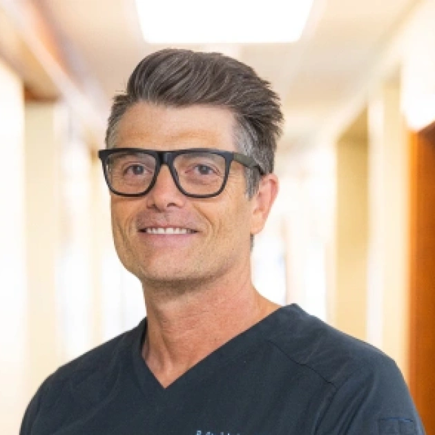 Professional man wearing glasses smiles in dental office hallway setting.